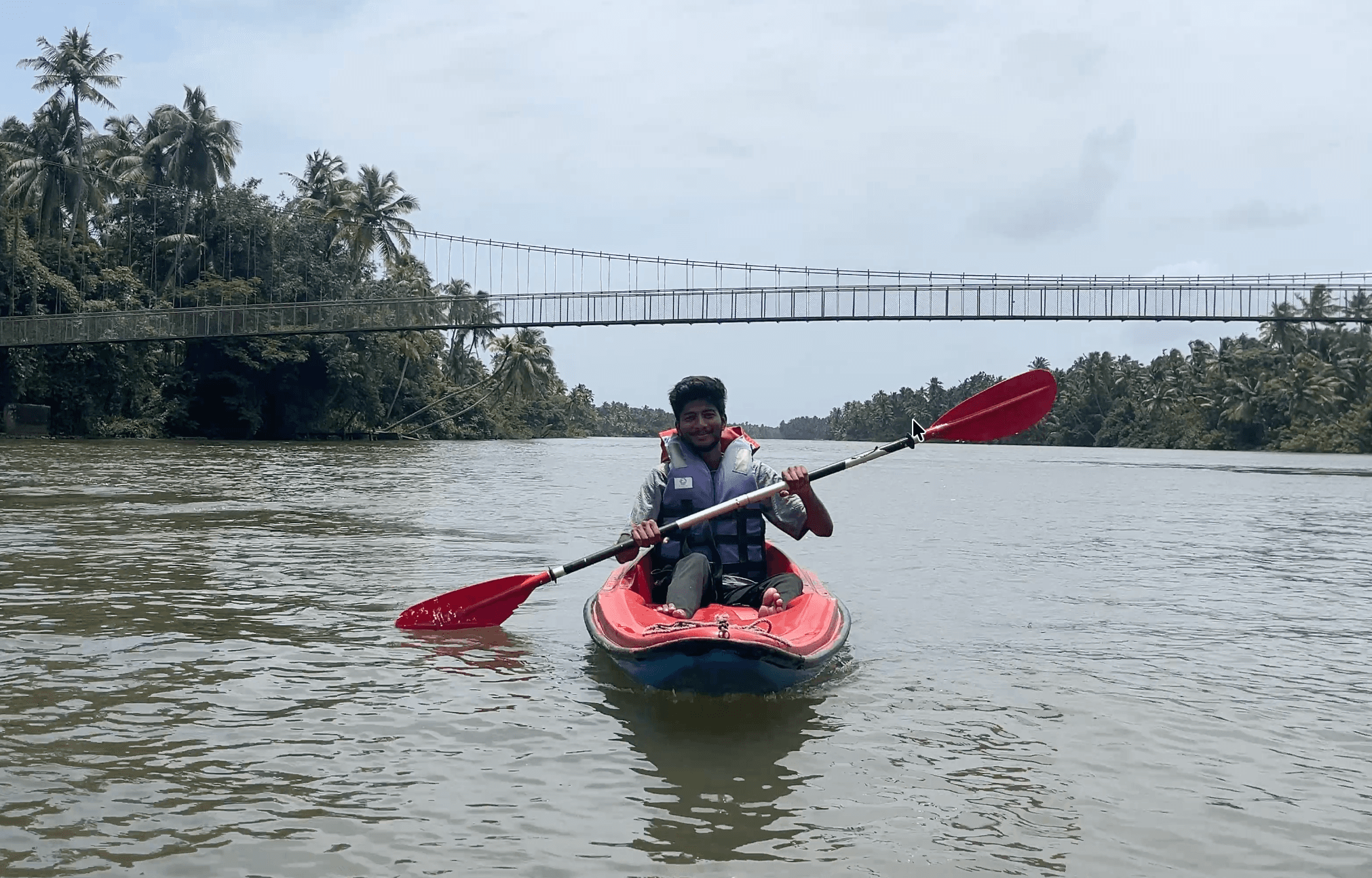 Tarak Ram kayaking on a calm river with lush green vegetation and a suspension bridge in the background